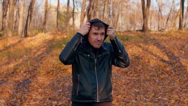 Mladý muž obléká kapotu v městském parku. Muž zahřívá hlavu v chladném počasí, zatímco stojí v podzimním lese. — Stock video