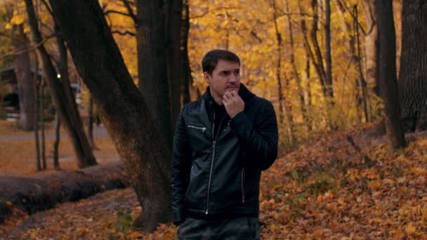 Joven mirando alrededor en el bosque de otoño. Varón pensativo se rasca la barbilla mientras está de pie en el parque de la ciudad. — Vídeos de Stock