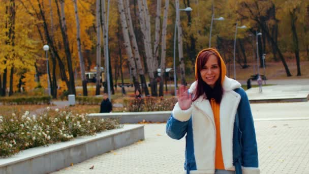 Mujer joven saluda, de pie en el parque de la ciudad. Hermosa hembra en ropa casual mirando a la cámara y sonriendo en la temporada de otoño. — Vídeos de Stock