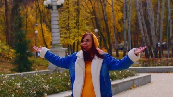 Young woman in city park. Beautiful female in casual clothes smiles, raising her hands, rejoicing in wind and fallen leaves in autumn season. — Stock Video