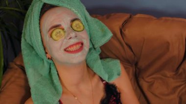 Yüzünde maske, gözlerinde salatalık dilimleri, koltukta dinlenen bir kadın. Yüzüne kozmetik maske takan rahat bir bayan. Kaplıca, cilt bakımı ve doğal güzellik kavramı..