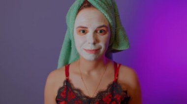 Başında havlu ve duştan sonra yüzünde maske olan genç bir kadın. Yüzüne makyaj maskesi takmış bir kadın. Kaplıca, cilt bakımı ve doğal güzellik kavramı..