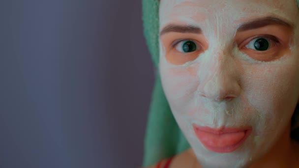 Donna con asciugamano sulla testa e maschera sul viso mostra lingua. Signora con maschera cosmetica viso guardando la fotocamera e flirtare. Concetto di spa, cura della pelle e bellezza naturale in condizioni domestiche. — Video Stock