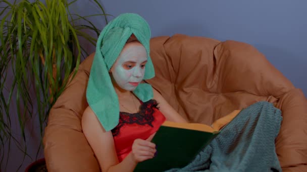 Kobieta z ręcznikiem na głowie, maską na twarzy, siedzi i czyta książki w fotelu. Pani dbająca o twarz maską kosmetyczną i odpoczywająca. Pojęcie spa, pielęgnacji skóry i naturalnego piękna w warunkach domowych. — Wideo stockowe