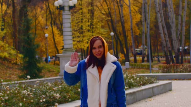 Mujer joven saluda, de pie en el parque de la ciudad. Hermosa hembra en ropa casual mirando a la cámara y sonriendo en la temporada de otoño. — Vídeos de Stock