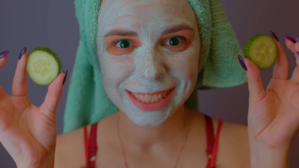 Jonge vrouw met handdoek op het hoofd en masker op het gezicht verbergen haar ogen achter komkommers en tonen hen. Lady verkering van haar gezicht met gezicht cosmetische masker en verse groenten. — Stockvideo
