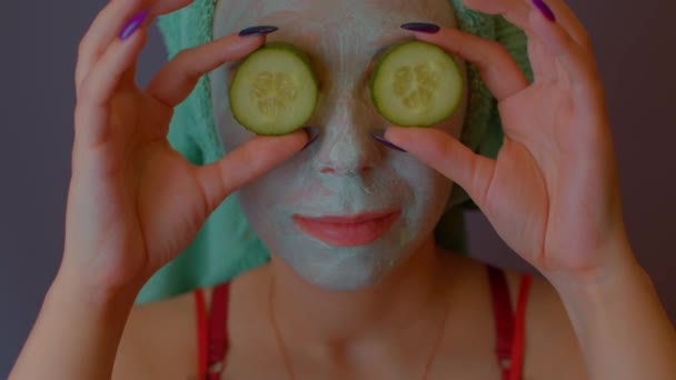 Junge Frau mit Handtuch auf dem Kopf und Maske im Gesicht versteckt ihre Augen hinter Gurken und zeigt sie. Lady umwirbt ihr Gesicht mit Kosmetikmaske und frischem Gemüse. — Stockvideo