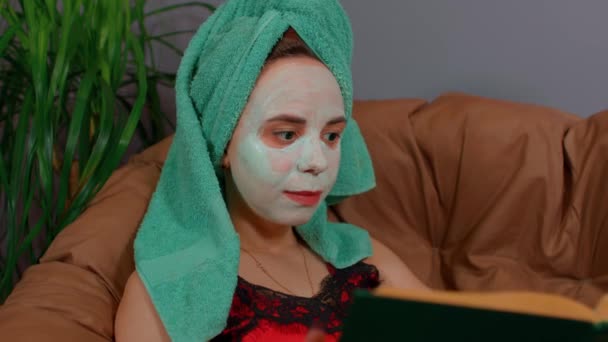 頭にタオル、顔にマスクをした女性が座り、アームチェアで本を読んでいます。化粧品マスクで顔を気にして休んでいる女性。国内の条件でスパ、スキンケア、自然の美しさの概念. — ストック動画