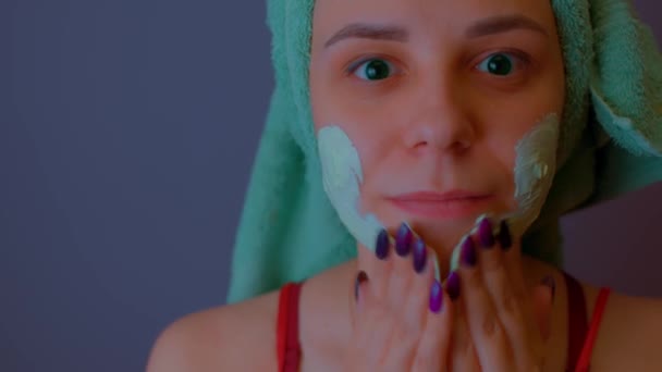 Young woman with towel on head applies mask on face after shower procedures. Lady courting of her face with facial cosmetic mask. Concept of spa, skincare and natural beauty in domestic conditions. — Stock Video