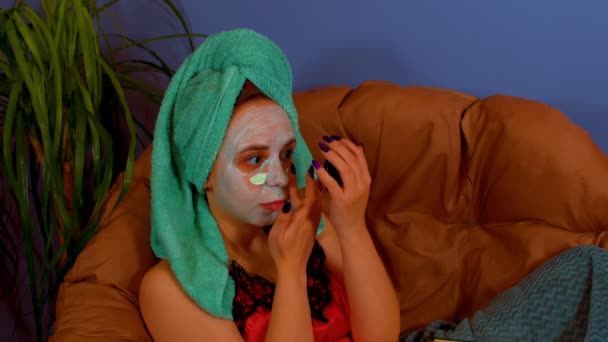 Νεαρή γυναίκα με πετσέτα στο κεφάλι εφαρμόζει μάσκα στο πρόσωπο, κάθεται στην πολυθρόνα. Η κυρία φροντίζει το πρόσωπό της με τη μάσκα προσώπου. Έννοια του σπα, της περιποίησης και της φυσικής ομορφιάς σε συνθήκες οικιακής χρήσης. — Αρχείο Βίντεο