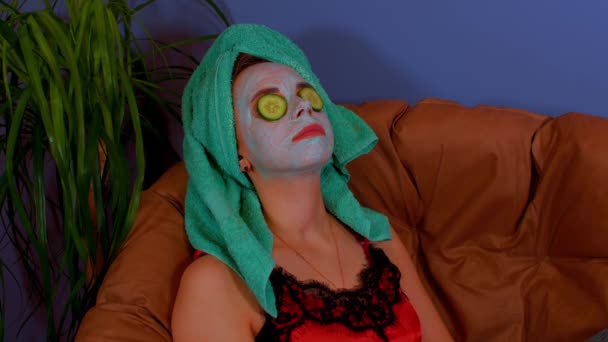 Vrouw met masker op het gezicht en komkommer plakjes op de ogen, rustend in een fauteuil. Ontspannen dame die voor haar gezicht zorgt met een cosmetisch masker. Begrip spa, huidverzorging en natuurlijke schoonheid in huiselijke omstandigheden. — Stockvideo