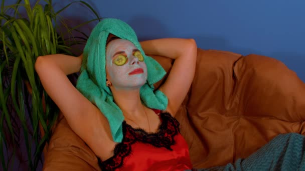 Vrouw met masker op het gezicht en komkommer plakjes op de ogen, rustend in een fauteuil. Ontspannen dame die voor haar gezicht zorgt met een cosmetisch masker. Begrip spa, huidverzorging en natuurlijke schoonheid in huiselijke omstandigheden. — Stockvideo