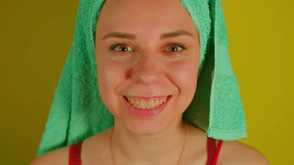 Een vrouw met een handdoek op haar hoofd en vlekken onder haar ogen glimlacht, kijkend naar de camera. Volwassen vrouw die haar gezicht het hof maakt in huiselijke omstandigheden. Concept van het bestrijden van rimpels en donkere kringen onder de ogen. — Stockfoto