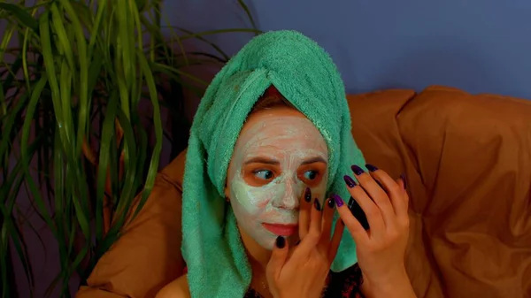 Ung kvinna med handduk på huvudet applicerar mask i ansiktet, sitter i fåtölj. Damen som bryr sig om ansiktet med kosmetisk ansiktsmask. Begreppet spa, hudvård och naturlig skönhet i hemmet. — Stockfoto
