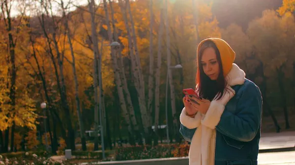 Junge Frau steht mit Handy im Stadtpark. Schöne Frau in lässiger Kleidung surft beim Spazierengehen im Smartphone. — Stockfoto