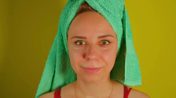 Woman with towel on head and patches under her eyes smiles, looking at camera. Adult female courting of her face in domestic conditions. Concept of fighting wrinkles and dark circles under eyes. — Stock Photo, Image
