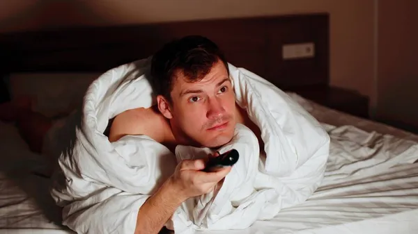 Junger Mann unter Decke auf dem Bett liegend und abends fernsehen. Entspanntes männliches Showprogramm und Kanalumschaltung mit Fernbedienung im dunklen Raum. — Stockfoto