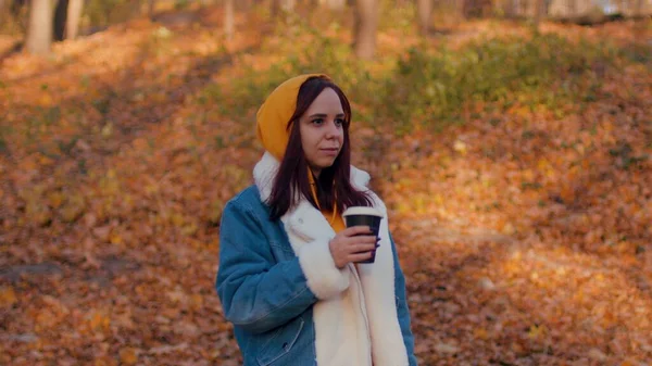 Jovem mulher bebendo café na floresta de outono. Mulher bonita em roupas casuais desfrutando de bebida quente em copo de papel em pé na floresta sazonal ensolarada. — Fotografia de Stock