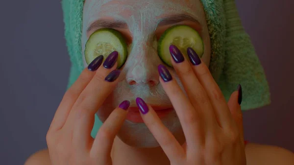 Frau mit Maske im Gesicht hält Gurkenscheiben auf den Augen. Entspannte Dame, die ihr Gesicht mit Kosmetikmaske und frischem Gemüse pflegt. Konzept von Wellness, Hautpflege und natürlicher Schönheit im häuslichen Umfeld. — Stockfoto