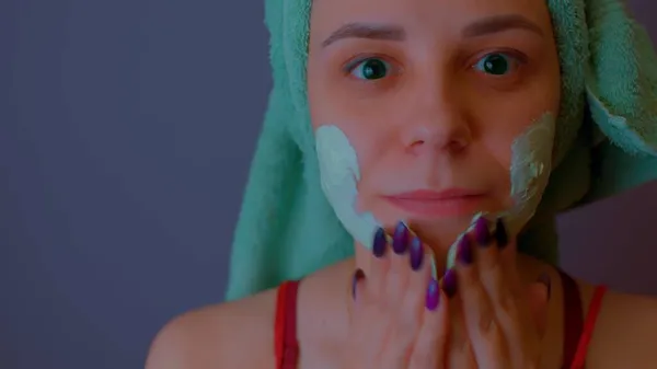 Ung kvinna med handduk på huvudet applicerar mask på ansiktet efter duschen förfaranden. Lady uppvaktning av hennes ansikte med ansiktskosmetisk mask. Begreppet spa, hudvård och naturlig skönhet i hemmet. — Stockfoto