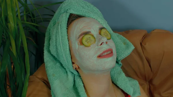 Egy nő maszkkal az arcán és uborka szeletekkel a szemén, karosszékben pihen. Nyugodt hölgy, aki kozmetikai maszkkal vigyáz az arcára. A gyógyfürdő, a bőrápolás és a természetes szépség fogalma hazai körülmények között. — Stock Fotó