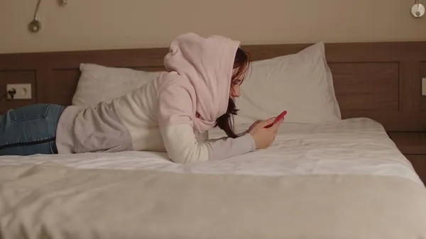 Junge Frau mit Kapuze und Handy auf dem Bett liegend. Entspannte Frau in Freizeitkleidung surft während der Pause im Smartphone. — Stockfoto