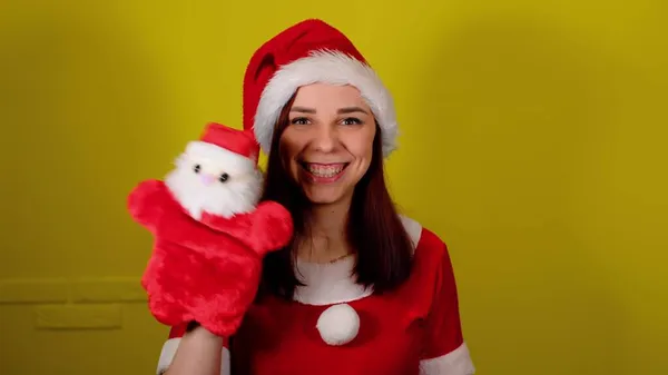 Žena v kostýmu Santy s měkkou loutkovou hračkou Santa Clause po ruce. Hezká fena se Santa Clausem loutkou na žlutém pozadí. Koncept loutkové výstavy a novoroční dovolené. — Stock fotografie