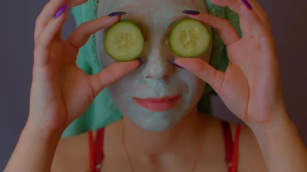 Young woman with towel on head and mask on face hiding her eyes behind cucumbers and showing them. Lady courting of her face with facial cosmetic mask and fresh vegetables. — Stock Photo, Image