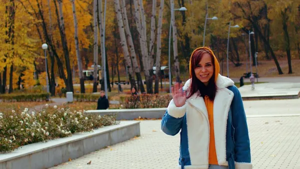 Mujer joven saluda, de pie en el parque de la ciudad. Hermosa hembra en ropa casual mirando a la cámara y sonriendo en la temporada de otoño. — Foto de Stock