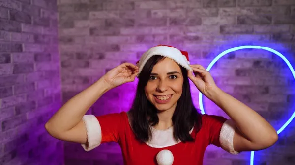 Noel Baba kostümü giymiş genç bir kadın Noel şapkasını takıyor. Güzel esmer, kameraya bakıyor ve aydınlanmış duvara gülümsüyor.. — Stok fotoğraf