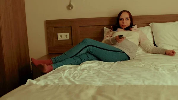 Młoda kobieta w luźnych ciuchach leżąca na poduszce w łóżku i oglądająca telewizję. Zmęczony kobiece programy oglądania show, przełącza kanał z pilotem. — Zdjęcie stockowe