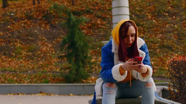 Giovane donna con cellulare nel parco cittadino. Bella femmina in abiti casual smartphone di navigazione mentre riposava sulla passeggiata. — Foto Stock