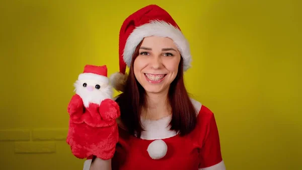 Donna in costume da Babbo Natale con peluche di Babbo Natale a portata di mano. Bella femmina con pupazzo di Babbo Natale su sfondo giallo. Concetto di spettacolo di marionette e vacanze di Capodanno. — Foto Stock