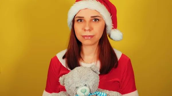 Télapó jelmezes nő, sárga hátterű macival. Csinos nő karácsonyi kalapban, plüss játékkal a kezében, kamerába néz és mosolyog.. — Stock Fotó