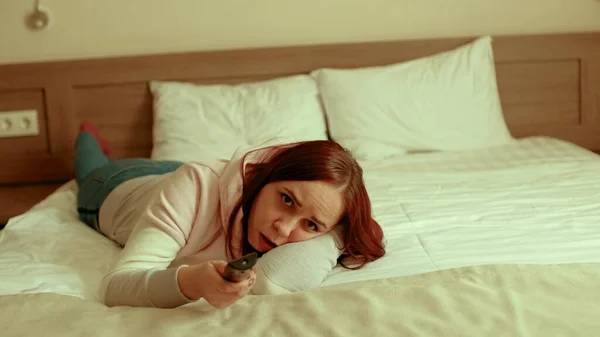 Młoda kobieta w luźnych ciuchach leżąca w łóżku i oglądająca telewizję. Zmęczone programy kobiece oglądania show, przełącza kanał z pilotem i zasypia. — Zdjęcie stockowe