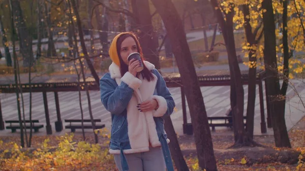 Jovem mulher bebendo café na floresta de outono. Mulher bonita em roupas casuais desfrutando de bebida quente em copo de papel em pé na floresta sazonal ensolarada. — Fotografia de Stock