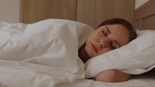 Ung kvinna som ligger på kudden under filten och sover på sängen på morgonen. Sömnig kvinna vaknar upp och ler, tittar på kameran. — Stockfoto