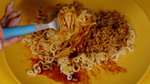 Nouilles instantanées prêtes à l'emploi avec condiment et sauce épicée dans une assiette jaune sur fond jaune. Personne méconnaissable avec des ramen mélangés fourchette avec des additifs. Gros plan. — Photo