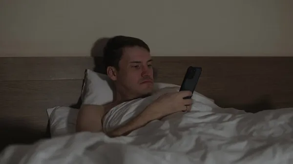 深夜赤身裸体的男人躺在枕头下，躺在床上，床上拿着手机。睡觉时放松男性浏览智能手机. 图库照片