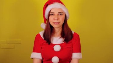 Sarı arka planda Noel Baba kostümlü bir kadın. Noel şapkalı güzel bir kadın kameraya bakıyor ve gülümsüyor..