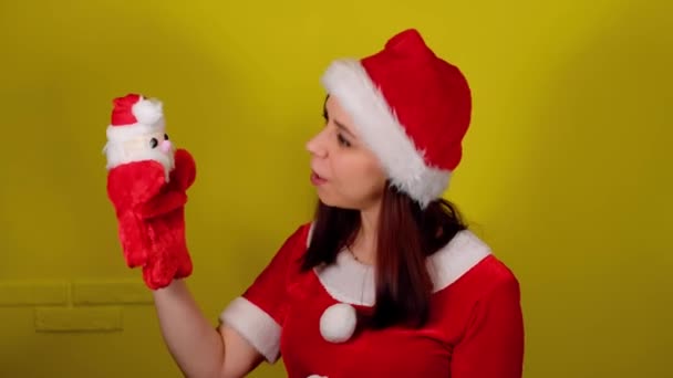 산타 복장을 한 여성 이 손에 쥐고 있는 산타 클로스의 부드러운 인형 장난감을 들고 있습니다. 노란 배경에 산타 클로스 인형 이 있는 예쁜 여자. 인형극의 개념 과 새해 휴일. — 비디오