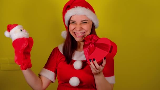 Жінка в костюмі Санти з м'якою маріонетковою іграшкою Санта-Клауса та подарунковою коробкою. Досить жіноча з маріонеткою Санта-Клауса і подарунком на жовтому тлі. Концепція маріонеткового шоу, новорічні свята та подарунки . — стокове відео