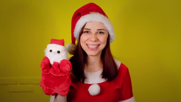 Frau im Weihnachtsmannkostüm mit Stoffpuppenspielzeug des Weihnachtsmannes an der Hand. Hübsches Weibchen mit Weihnachtsmann-Puppe auf gelbem Hintergrund. Konzept für Puppenspiel und Neujahrsferien. — Stockvideo