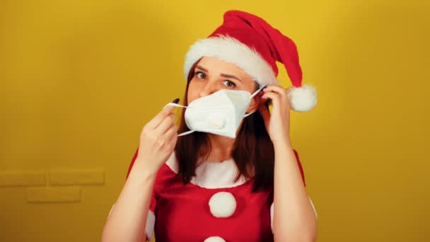 Mujer en traje de santa pone máscara médica en la cara. Bastante hembra en sombrero de Navidad pone máscara protectora, mirando a la cámara. — Vídeo de stock