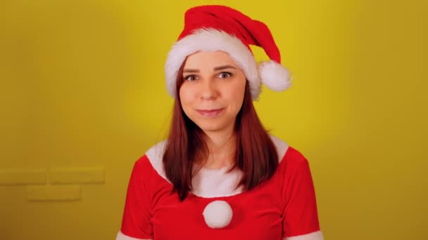 Woman in santa costume winks on yellow background. Pretty female in christmas hat flirting and looking at camera. — Stock Video