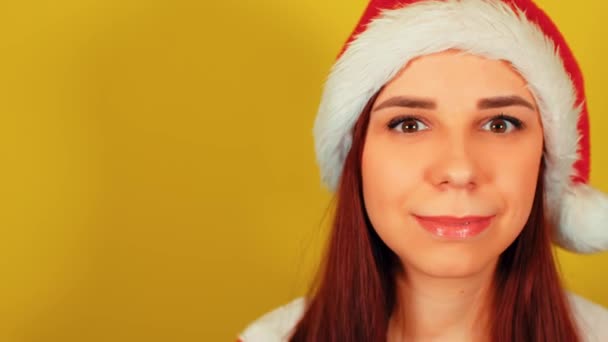 サンタの衣装の女性は黄色の背景でウィンクします。かわいいです女性でクリスマス帽子イチャイチャと見ますカメラ. — ストック動画
