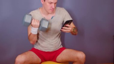 Spor bantlı bir adam dambılı kaldırıyor, cep telefonuna bakıyor ve jimnastik topunun üzerinde oturuyor. Aktif erkek spor yapıyor ve eğleniyor, fitness topunun üzerinde oturuyor..