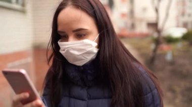 Yüzünde tıp maskesi olan genç bir kadın sokaklarda cep telefonundan salgın hakkında haberler öğreniyor. Yetişkin kadın kendini hastalıklardan korumak için yüzünü maskeyle örttü..