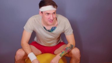 Spor bantlı bir adam jimnastik topunda zıplıyor. Aktif erkek evde fitness topuyla ısınıyor..