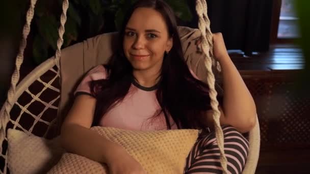 快乐的年轻女人在家里的吊床上休息 快乐的年轻貌美的女性坐在家里的吊床上 一边看着镜头一边看着镜头 — 图库视频影像